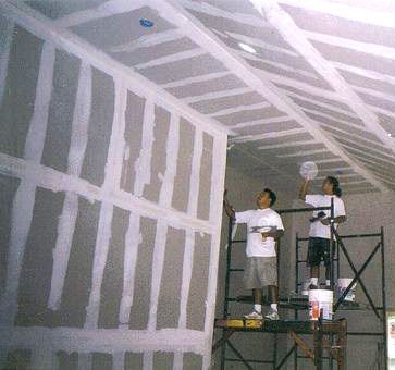 Dos personas en un andamio, colocando paneles de yeso en las paredes y el techo de una habitación. Se ven el compuesto blanco y las paredes grises.