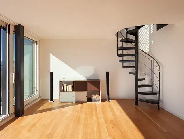 Luminoso salón con suelo de madera, escalera de caracol y gran ventanal que proyecta sombras.