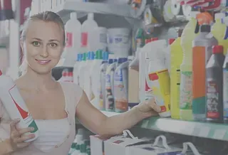 Mujer sonriendo en una ferretería, sosteniendo sellador y alcanzando un producto de limpieza en el estante.