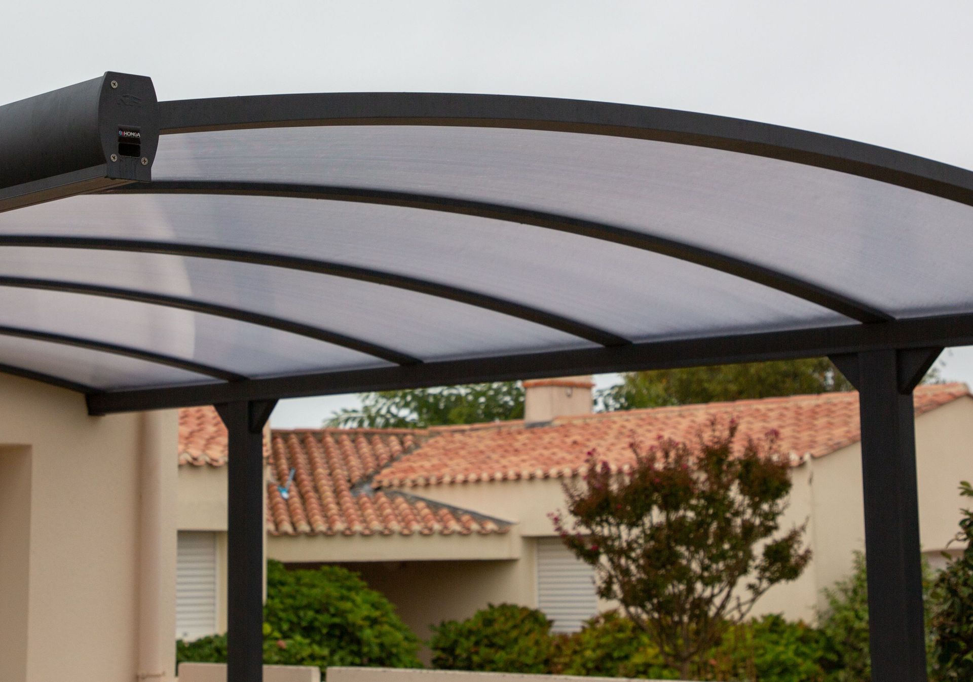 Pérgola de metal negro con soportes arqueados y techo translúcido, con vistas a una casa con tejas naranjas.