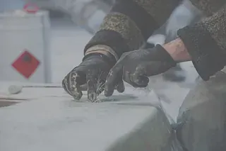 Manos enguantadas de una persona trabajando con material polvoriento; en el fondo hay un contenedor con un símbolo de peligro.