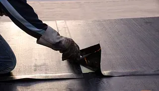 Una persona con guantes trabaja en un techo oscuro, doblando un trozo de material cerca de una costura.