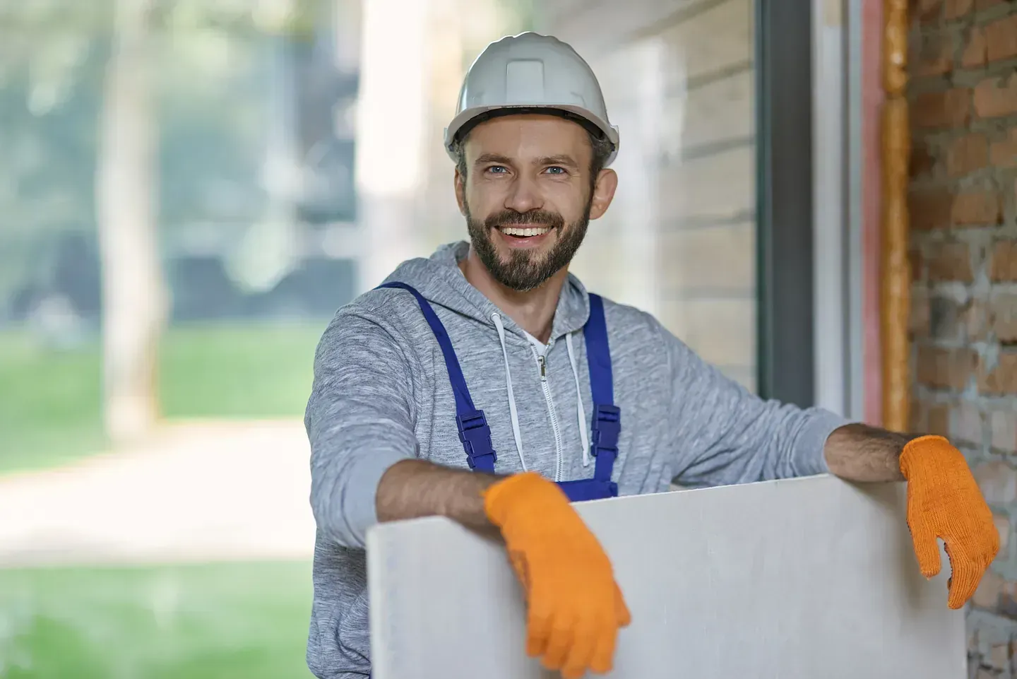 Obrero de la construcción con casco, mono, guantes naranjas, sonriendo, sosteniendo placas de yeso.