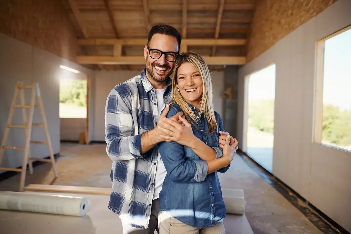 Pareja abrazándose y sonriendo en una casa sin terminar, abrazados; expresiones felices.
