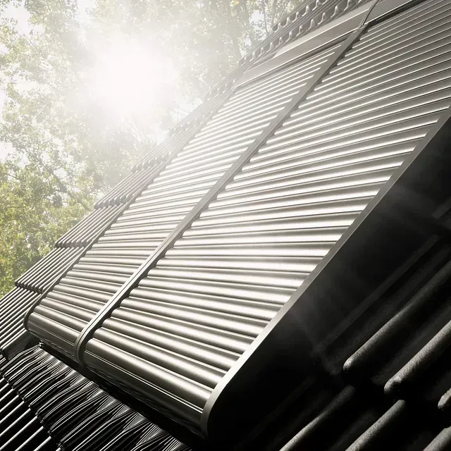 Contraventanas metálicas, de lamas, parcialmente cerradas, en ángulo contra un fondo borroso de árboles y luz solar.