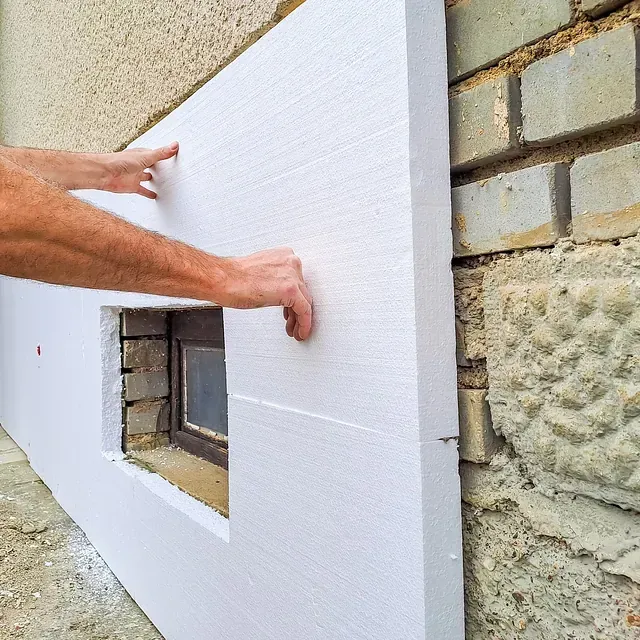 Una persona instalando aislamiento de espuma blanca en una pared exterior de ladrillo, alrededor de una ventana pequeña.