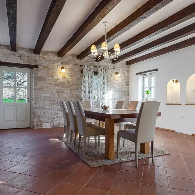 Comedor con vigas de madera, paredes de piedra, mesa puesta para comer y vista a un jardín.