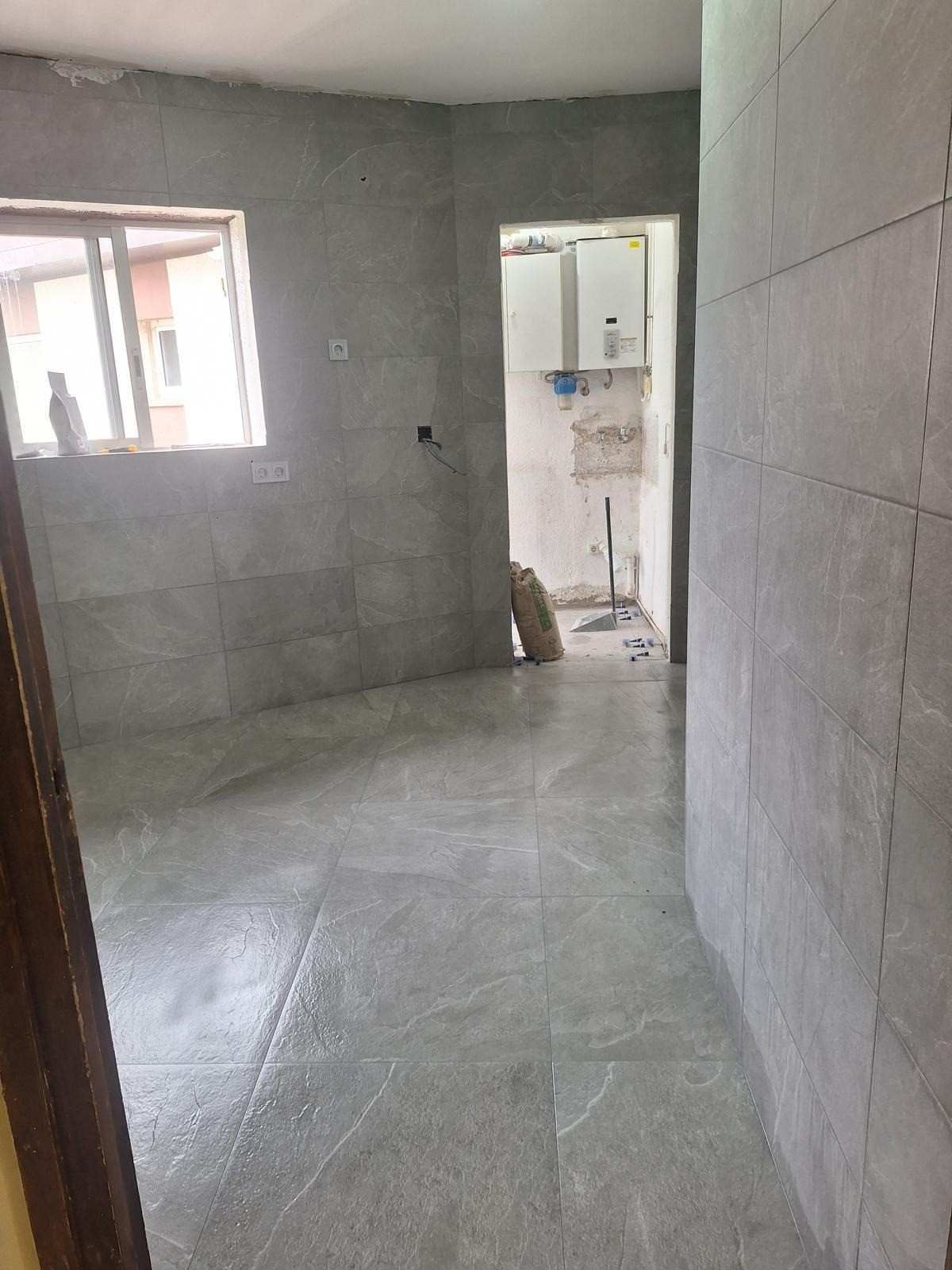 Habitación vacía en construcción con azulejos, paredes grises, una ventana y una puerta que da a un pequeño baño.