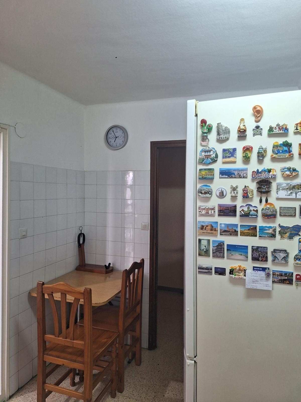 Pequeña cocina con mesa y sillas de madera, reloj de pared y nevera cubierta de imanes y fotos.