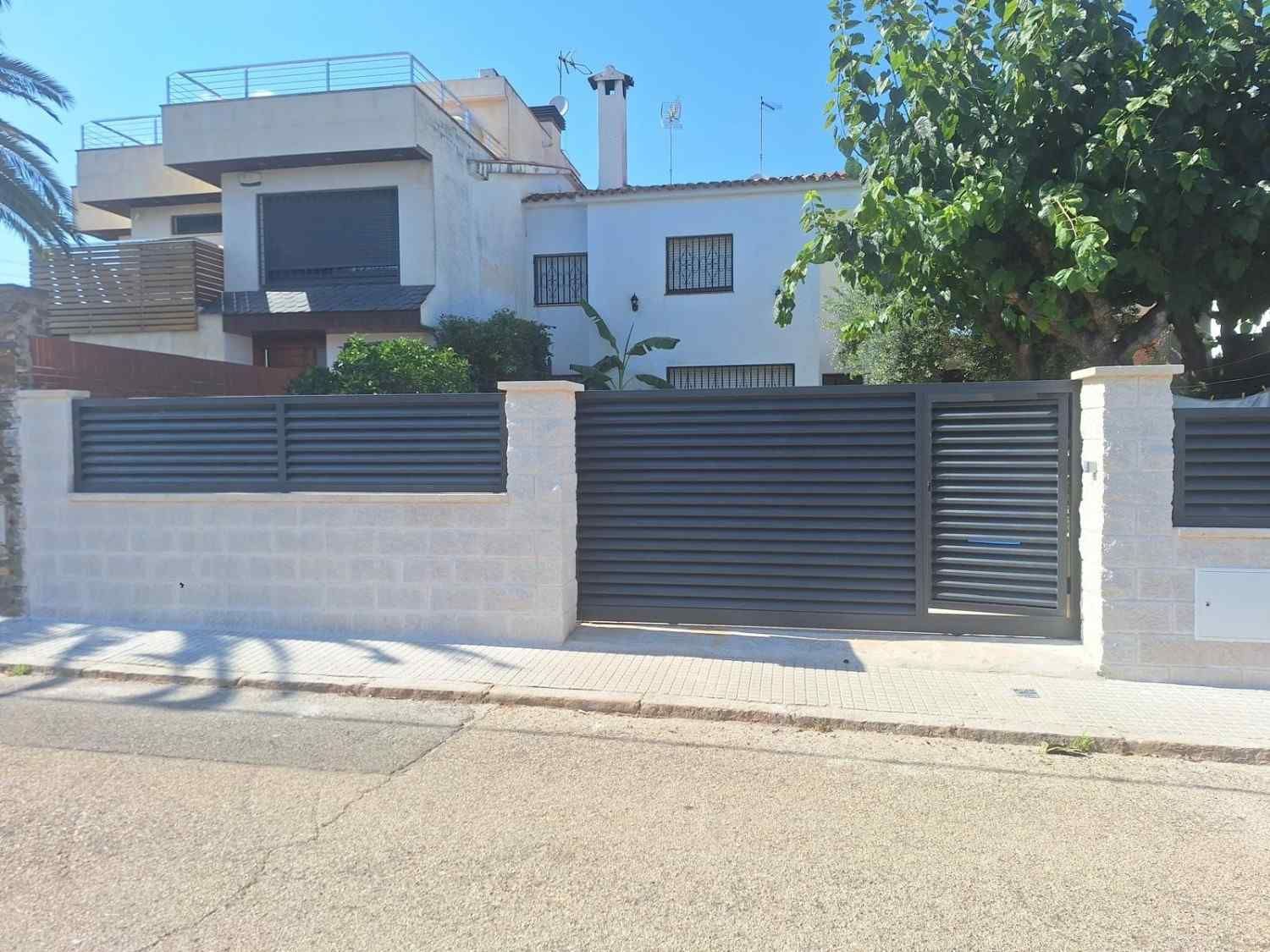 Casa moderna detrás de un muro blanco y una puerta gris oscuro en una calle soleada.