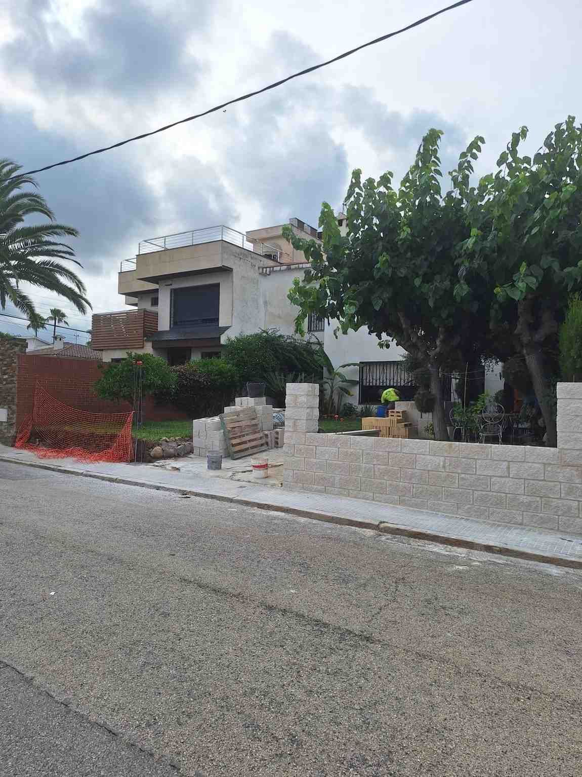 Calle residencial con una casa blanca, un árbol frondoso y un muro bajo de piedra bajo un cielo nublado.