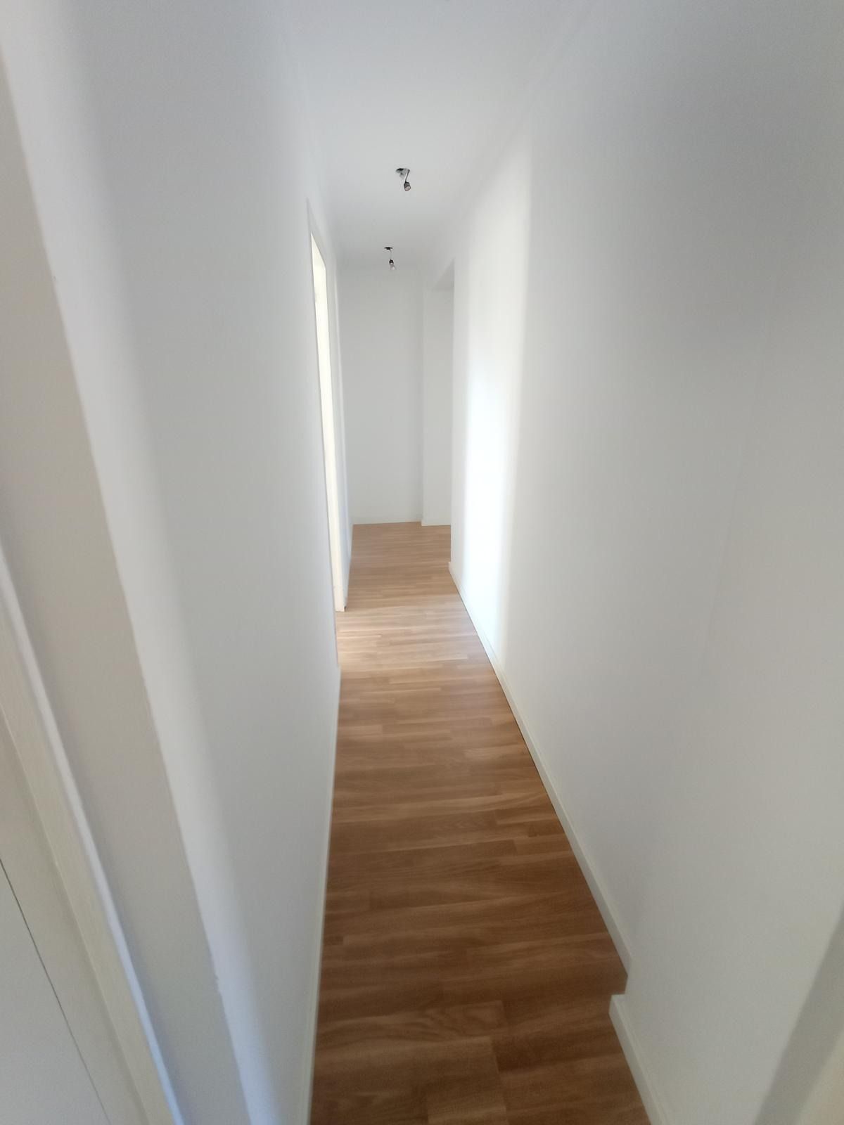 Pasillo estrecho y blanco con suelo de madera y luz brillante al final.