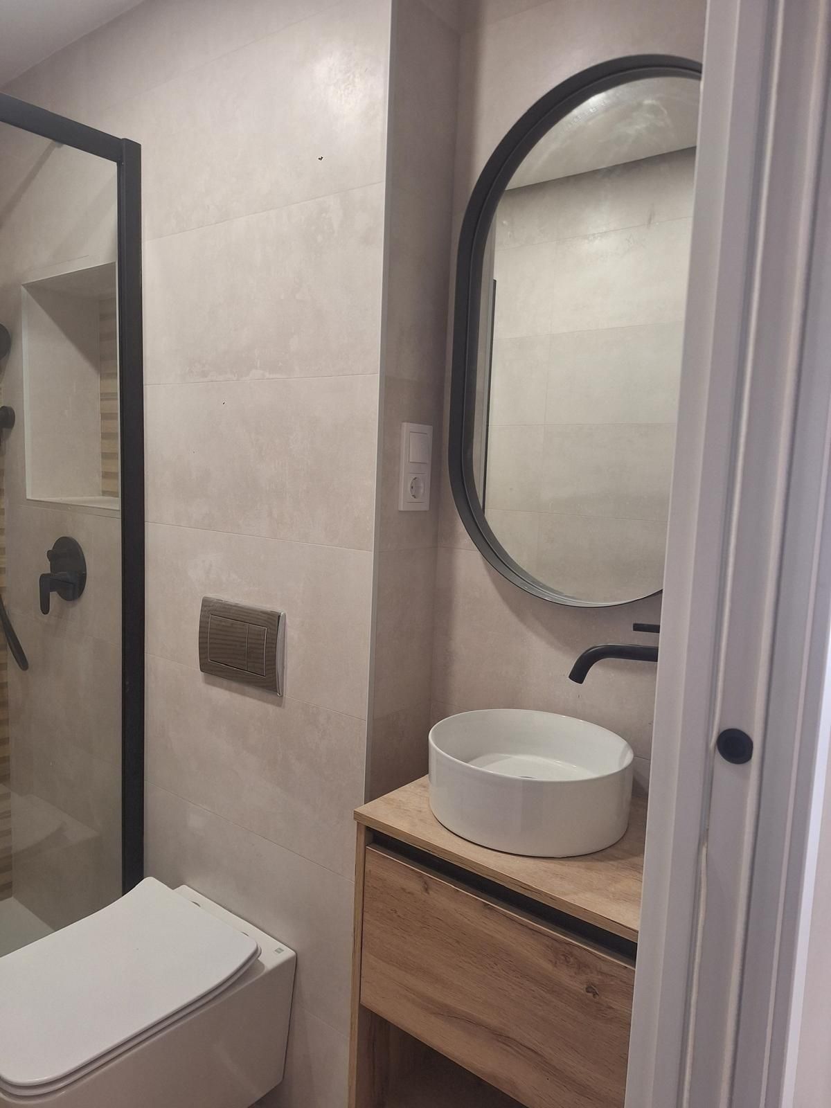 Baño con inodoro blanco, lavabo de madera, espejo ovalado y ducha visible a la izquierda.