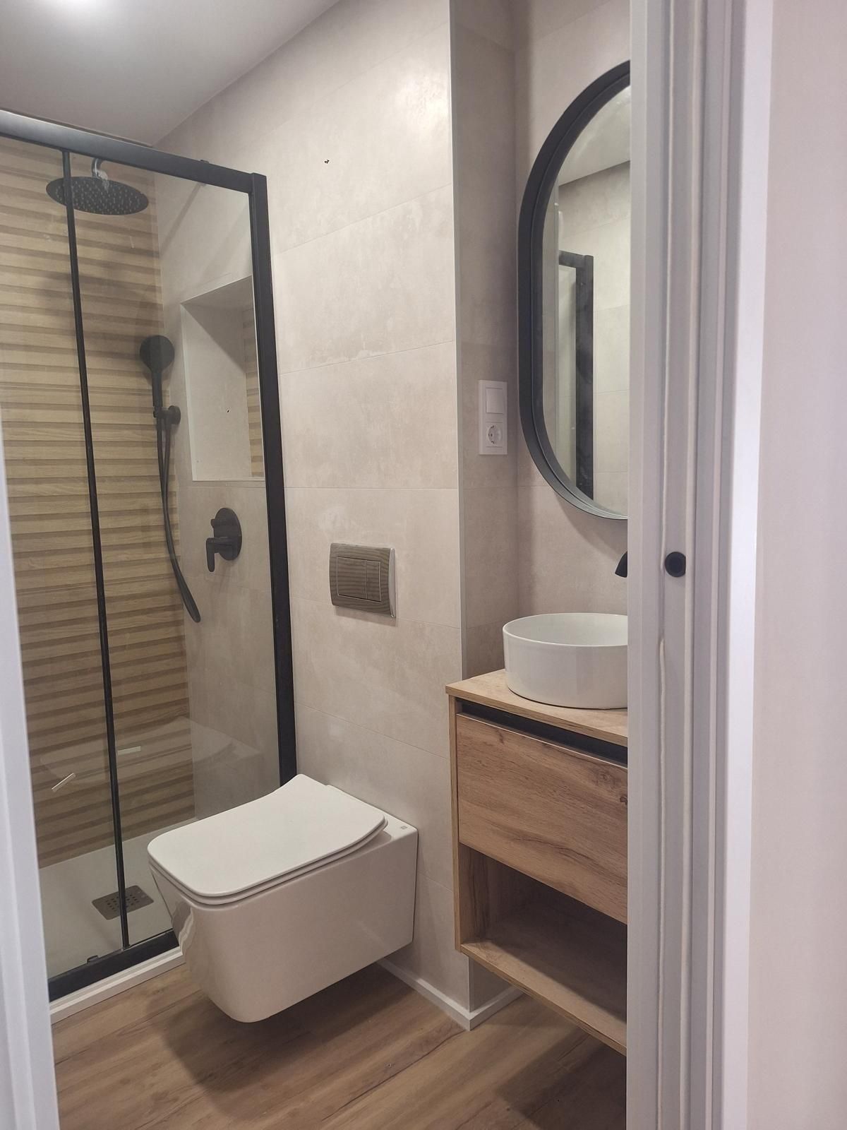Baño moderno con ducha de cristal, inodoro suspendido y tocador de madera con espejo redondo.