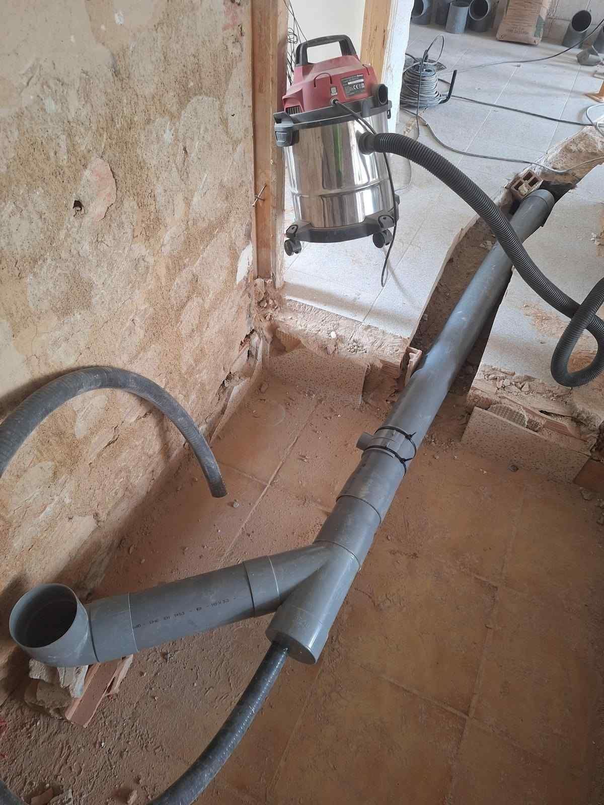 Tuberías de PVC grises y una manguera de aspiradora húmeda sobre un suelo de baldosas junto a una pared, lo que sugiere trabajos de fontanería o limpieza.