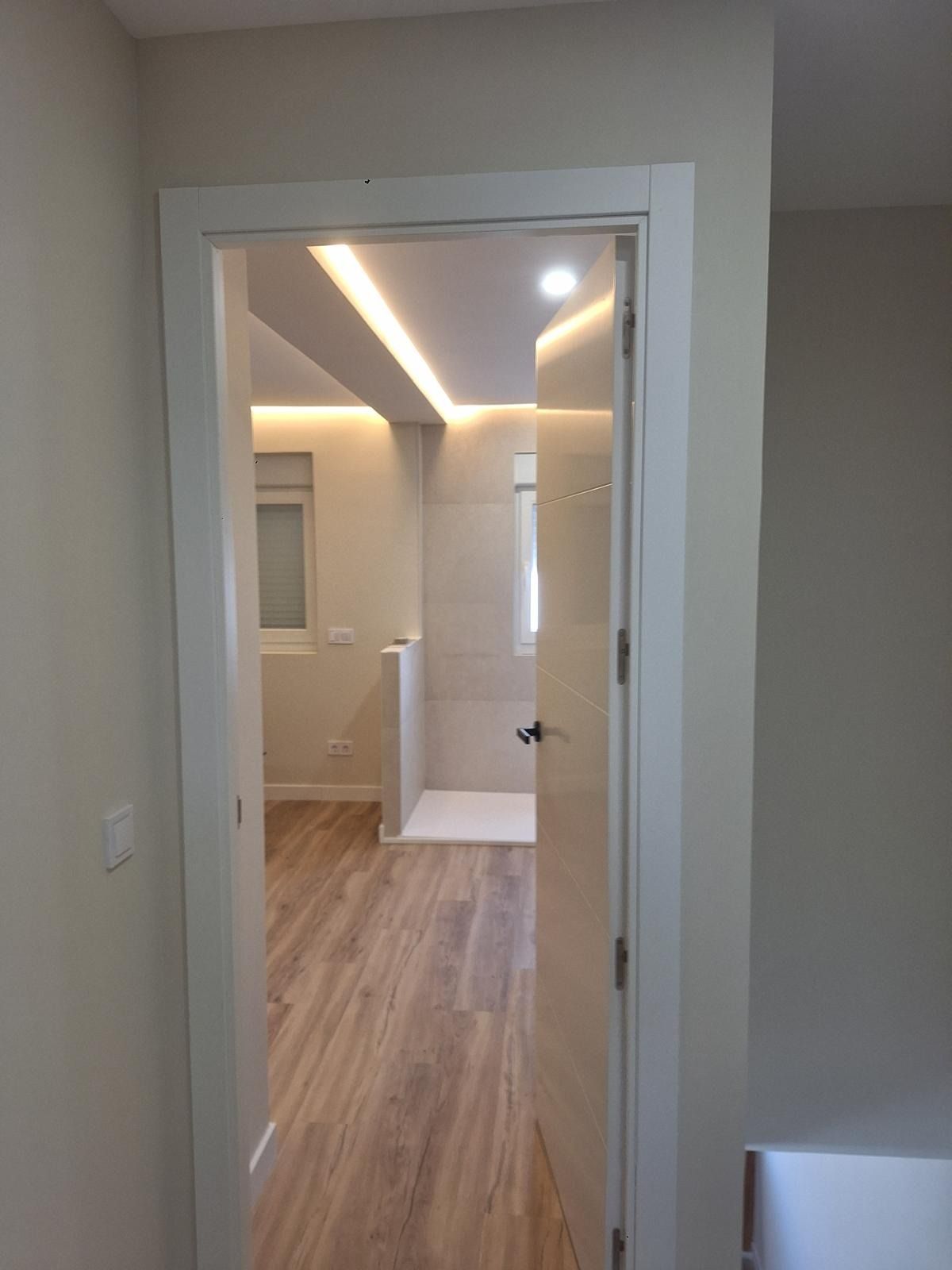 Pasillo blanco que conduce a una luminosa habitación moderna con suelo de madera y luces empotradas en el techo.