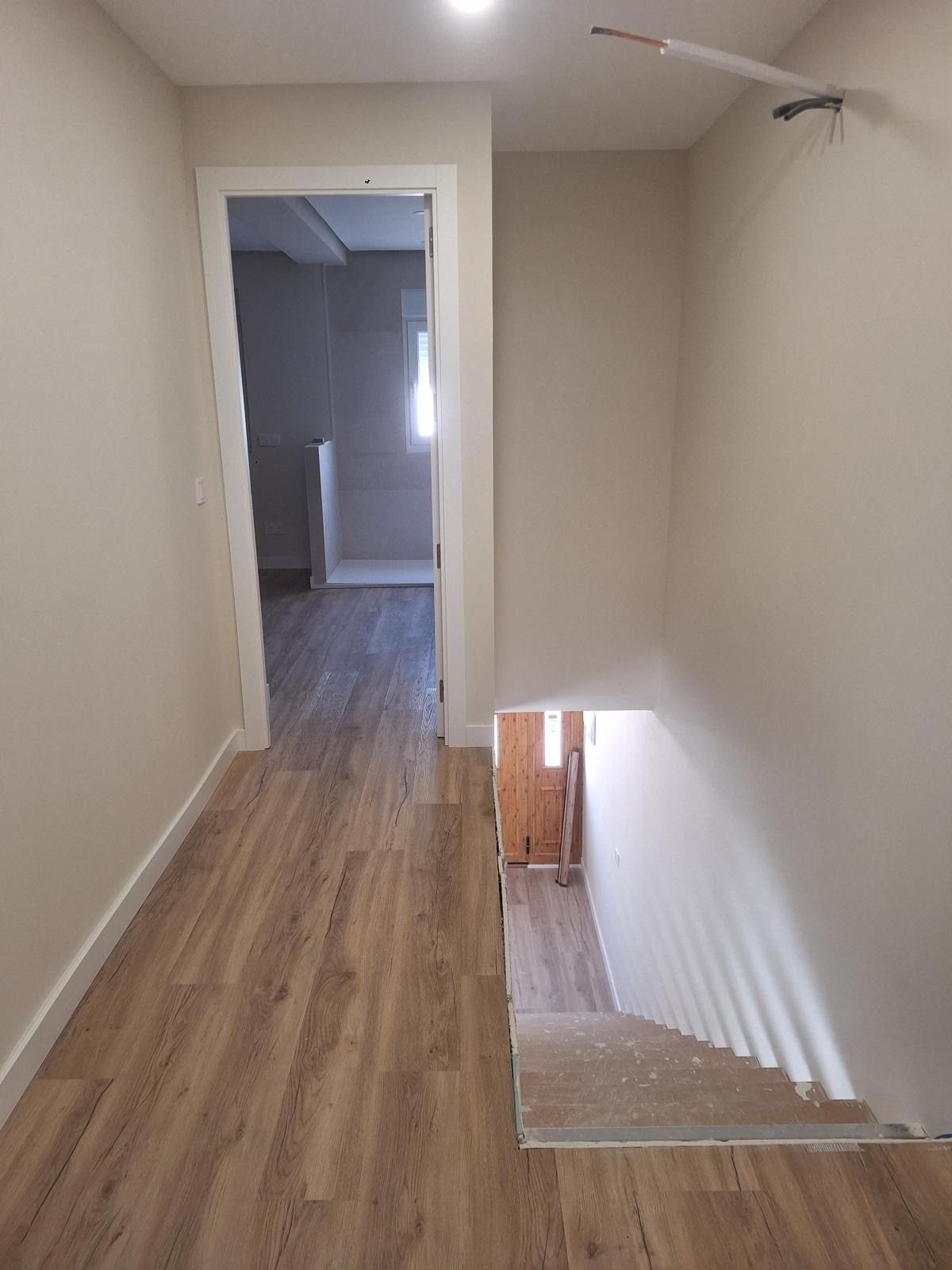 Pasillo vacío con suelo de madera y una abertura de escalera a la derecha, iluminado por luz natural.