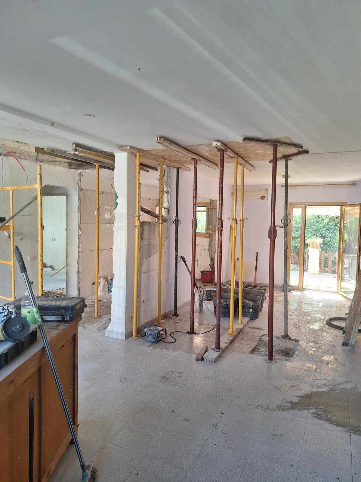 Interior en proceso de renovación con vigas de pared a la vista, herramientas y suelo sin terminar, iluminado por la luz natural que entra por las puertas abiertas.