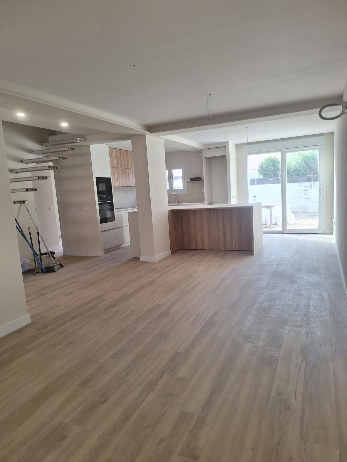 Sala de estar moderna y vacía con pisos de madera clara, paredes blancas, escalera e isla de cocina junto a puertas de cristal.