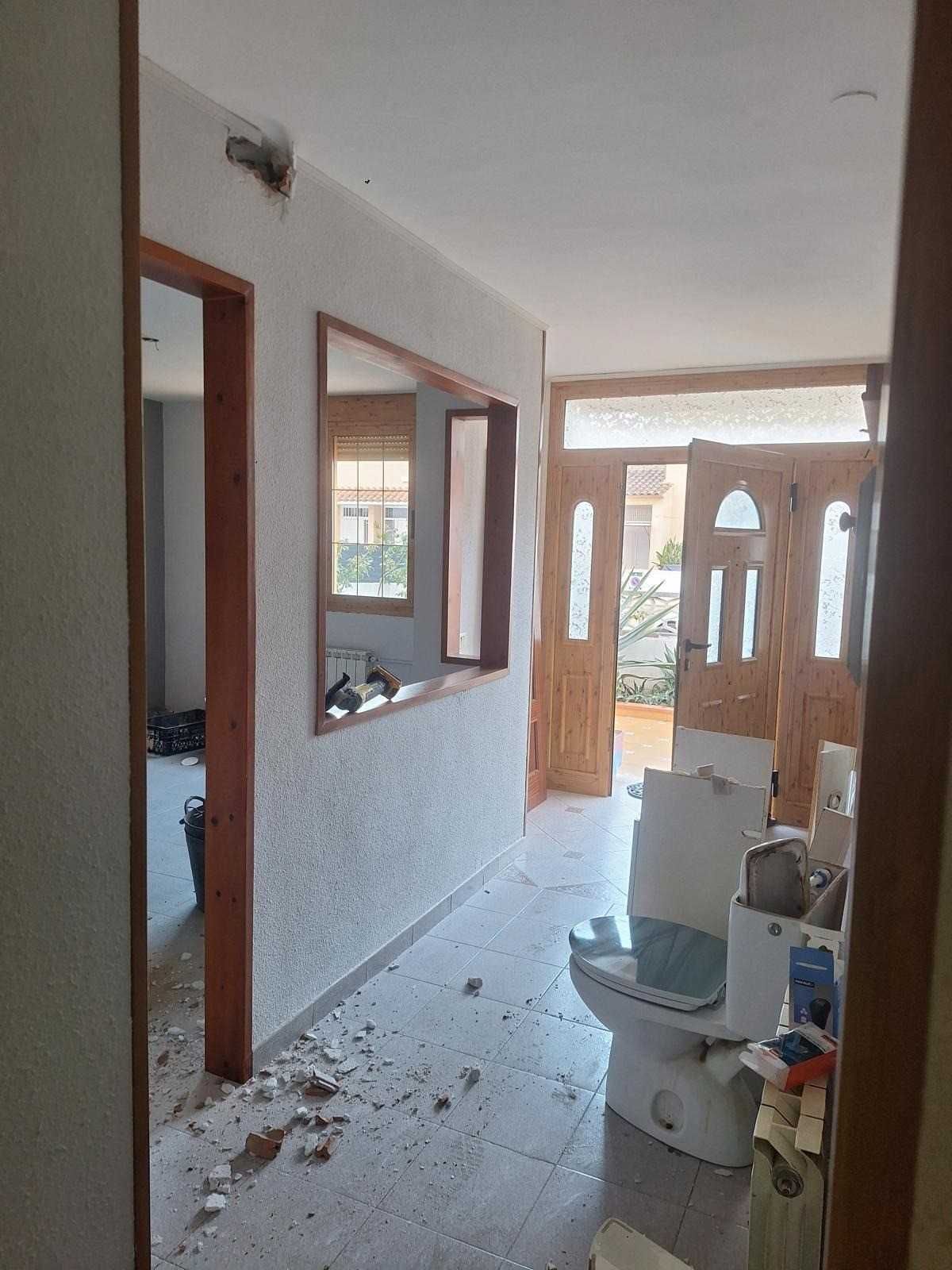 Pasillo estrecho en obras, con escombros en el suelo, puertas abiertas y un inodoro cerca de la cámara.