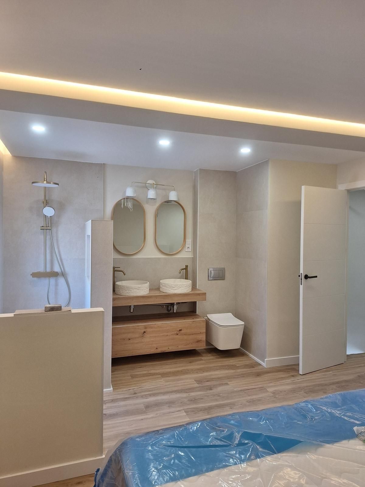 Baño moderno con lavabos dobles, espejos, ducha e inodoro; suelo parcialmente cubierto por reformas.