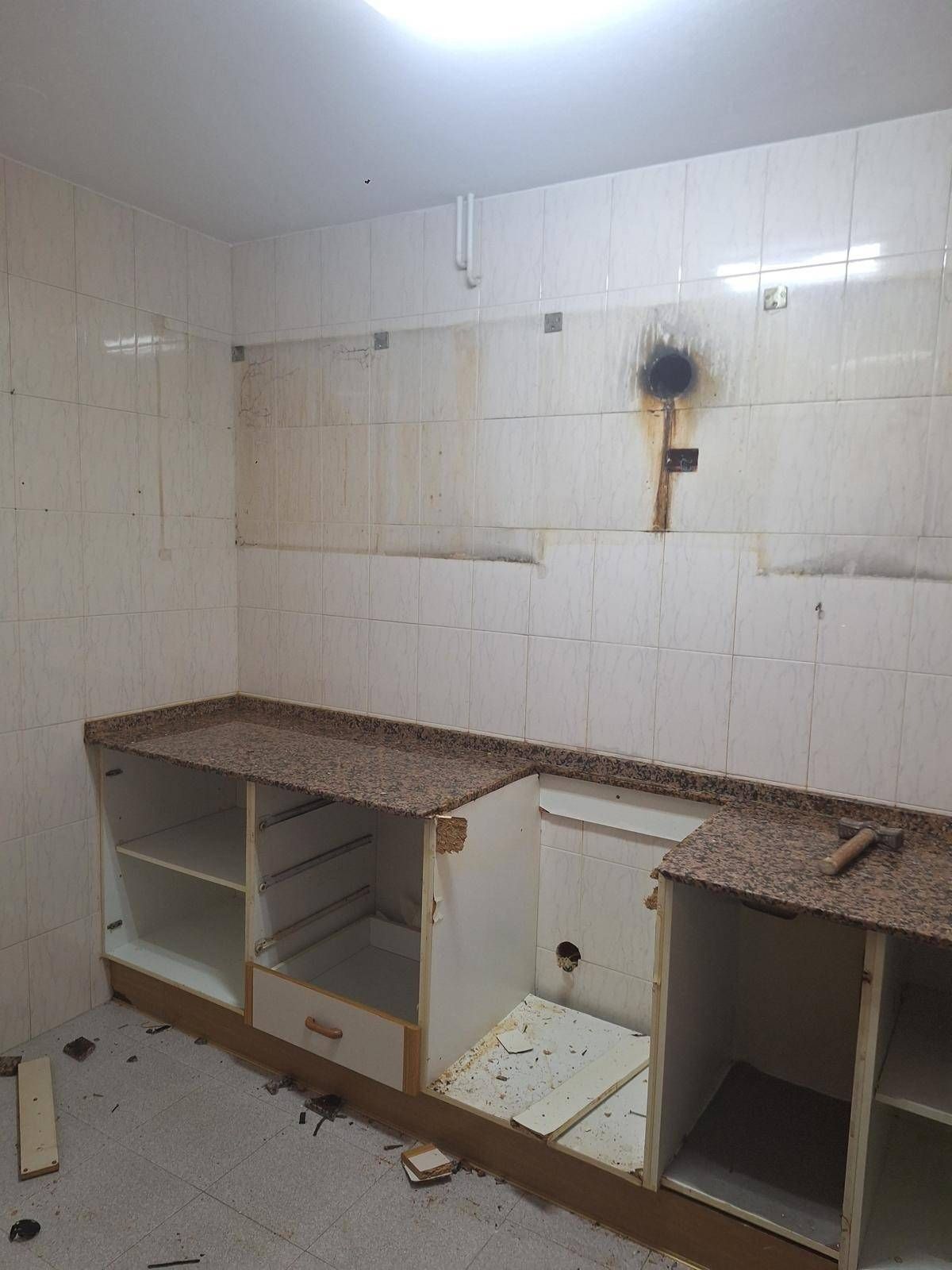 Cocina dañada con paredes de azulejos blancos rotos, armarios faltantes y escombros en el suelo.
