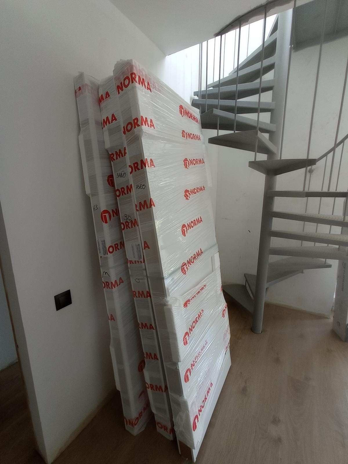 Colchones apilados envueltos en plástico junto a una escalera de caracol metálica en una habitación luminosa.