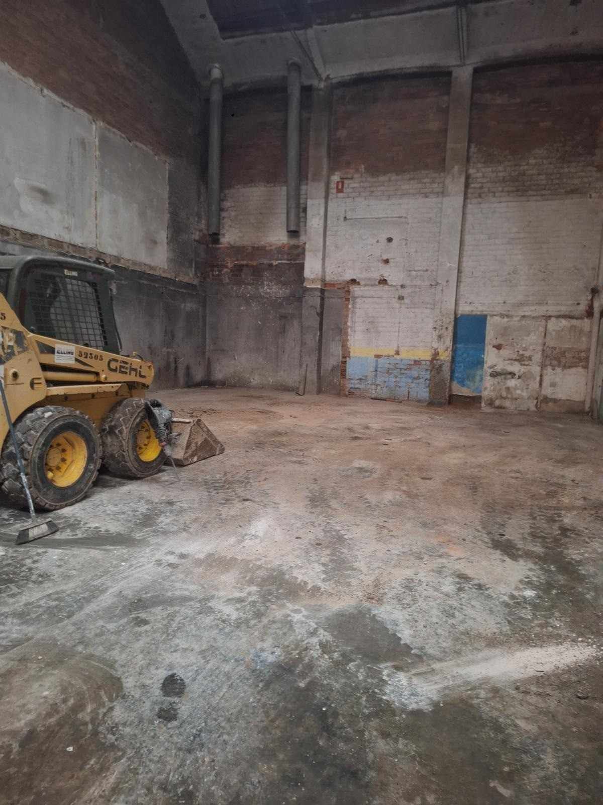 Interior vacío de un almacén con una minicargadora amarilla a la izquierda y paredes de hormigón.