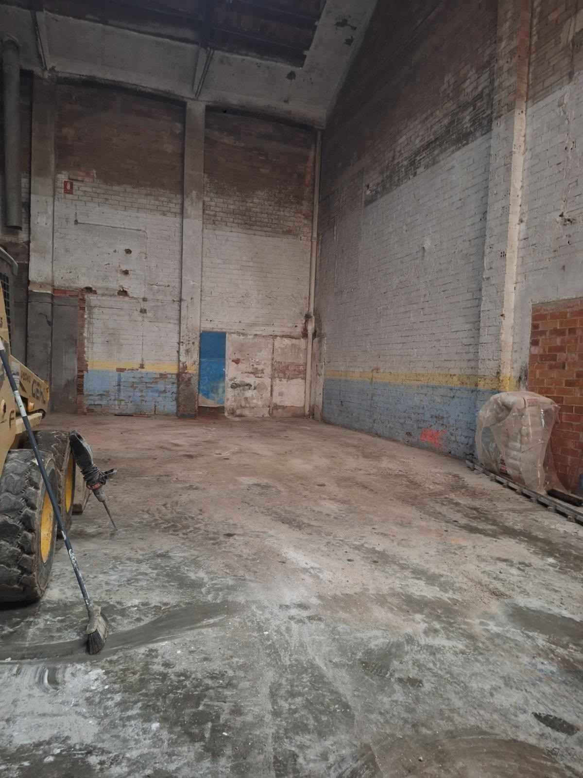Nave industrial vacía con suelo de hormigón, paredes de ladrillo blanco y una máquina amarilla en el extremo izquierdo.