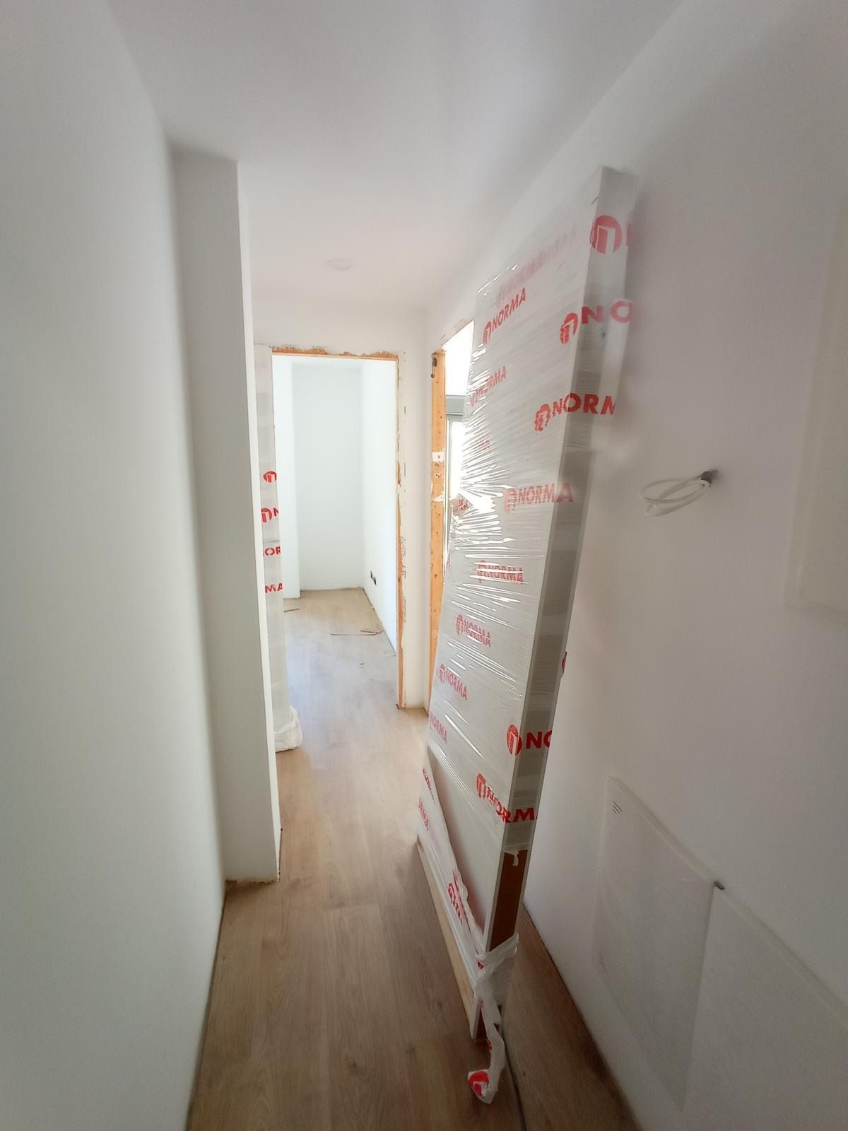 Pasillo estrecho en obras de renovación con marco de puerta revestido, paredes blancas y suelo de madera.