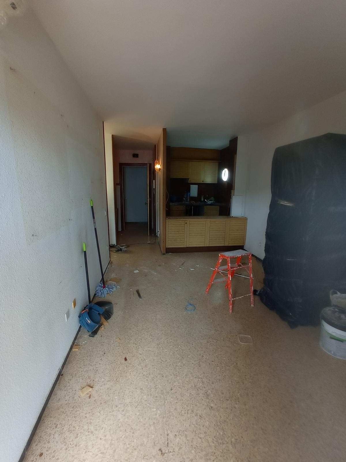 Pasillo estrecho con escombros esparcidos, un taburete rojo, un gran armario negro y una zona de cocina al final.