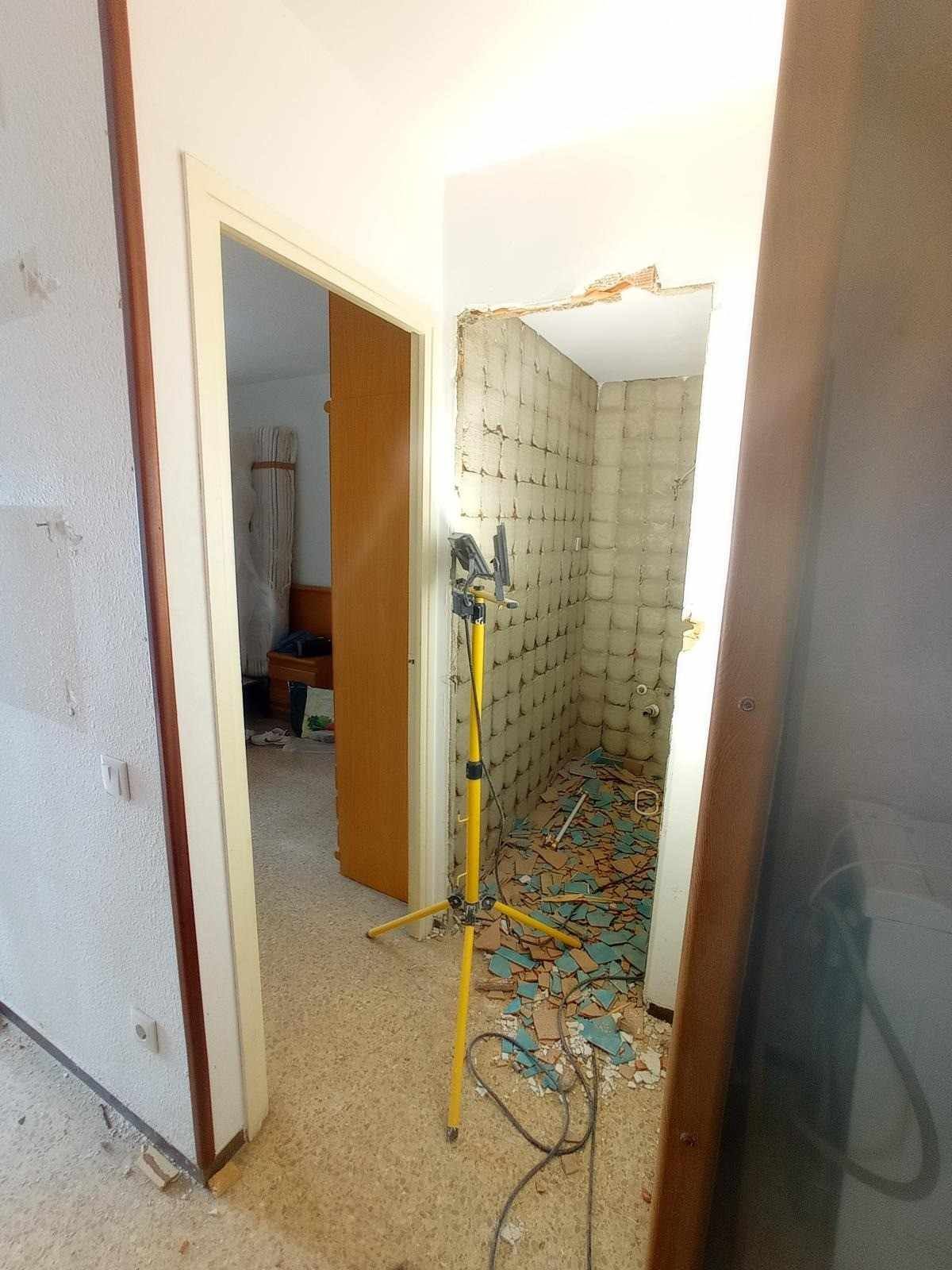 Entrada de baño dañada con escombros y cableado expuesto en un pasillo estrecho.