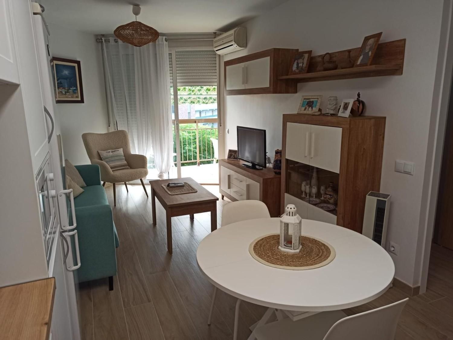 Sala de estar luminosa con mesa redonda blanca, sofá color turquesa, mueble para la televisión y puertas que dan al balcón.