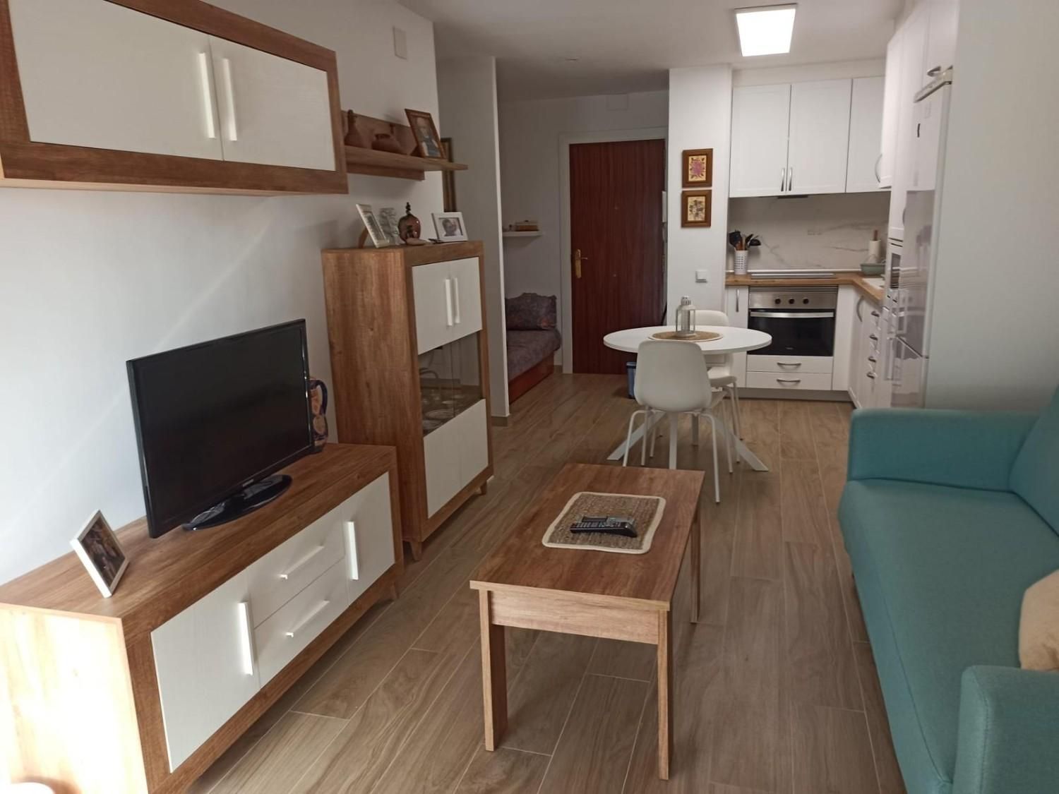 Pequeña sala de estar con televisor, mesa de centro, sofá color turquesa y cocina abierta al fondo.