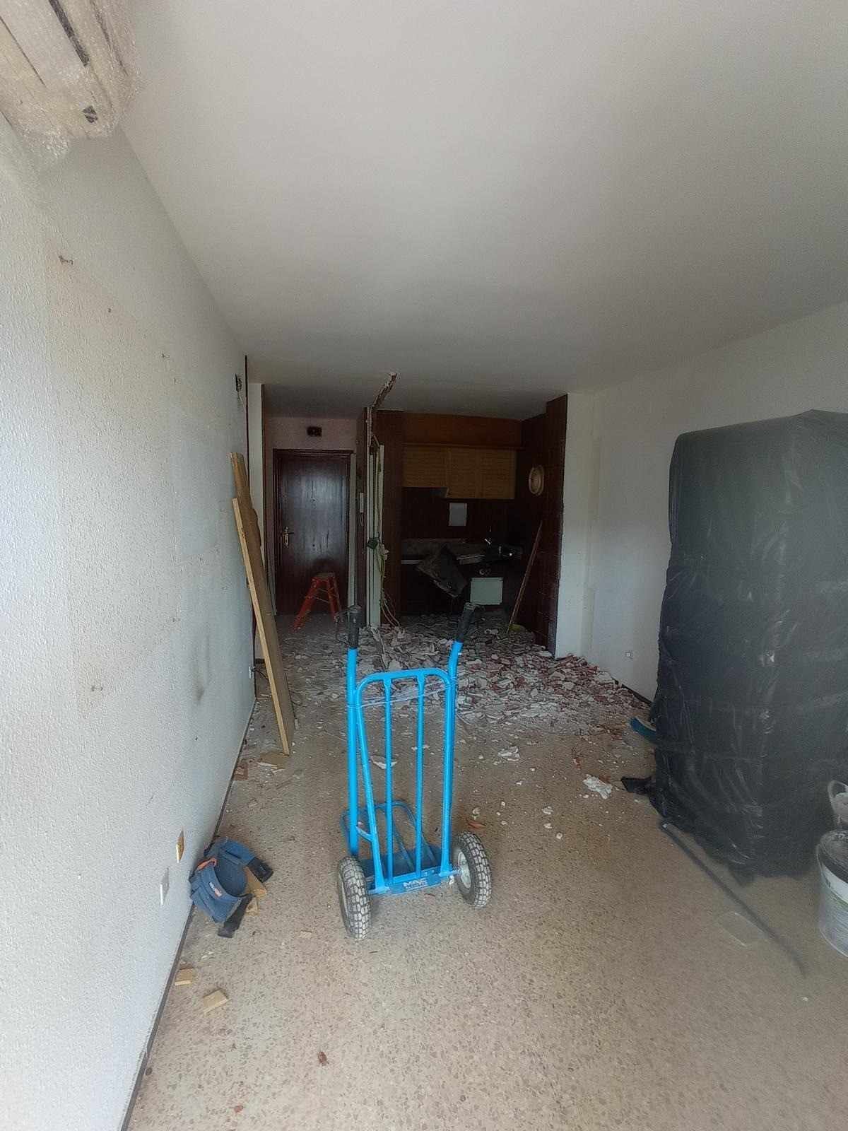 Un pasillo estrecho y desordenado, lleno de escombros, una carretilla azul en el centro y una habitación parcialmente abierta al final.