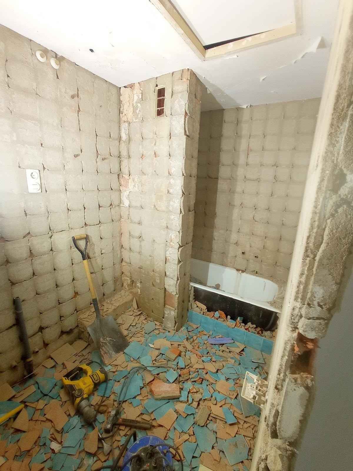 Interior del baño dañado, con baldosas azules rotas en el suelo, paredes desconchadas y escombros esparcidos por todas partes.