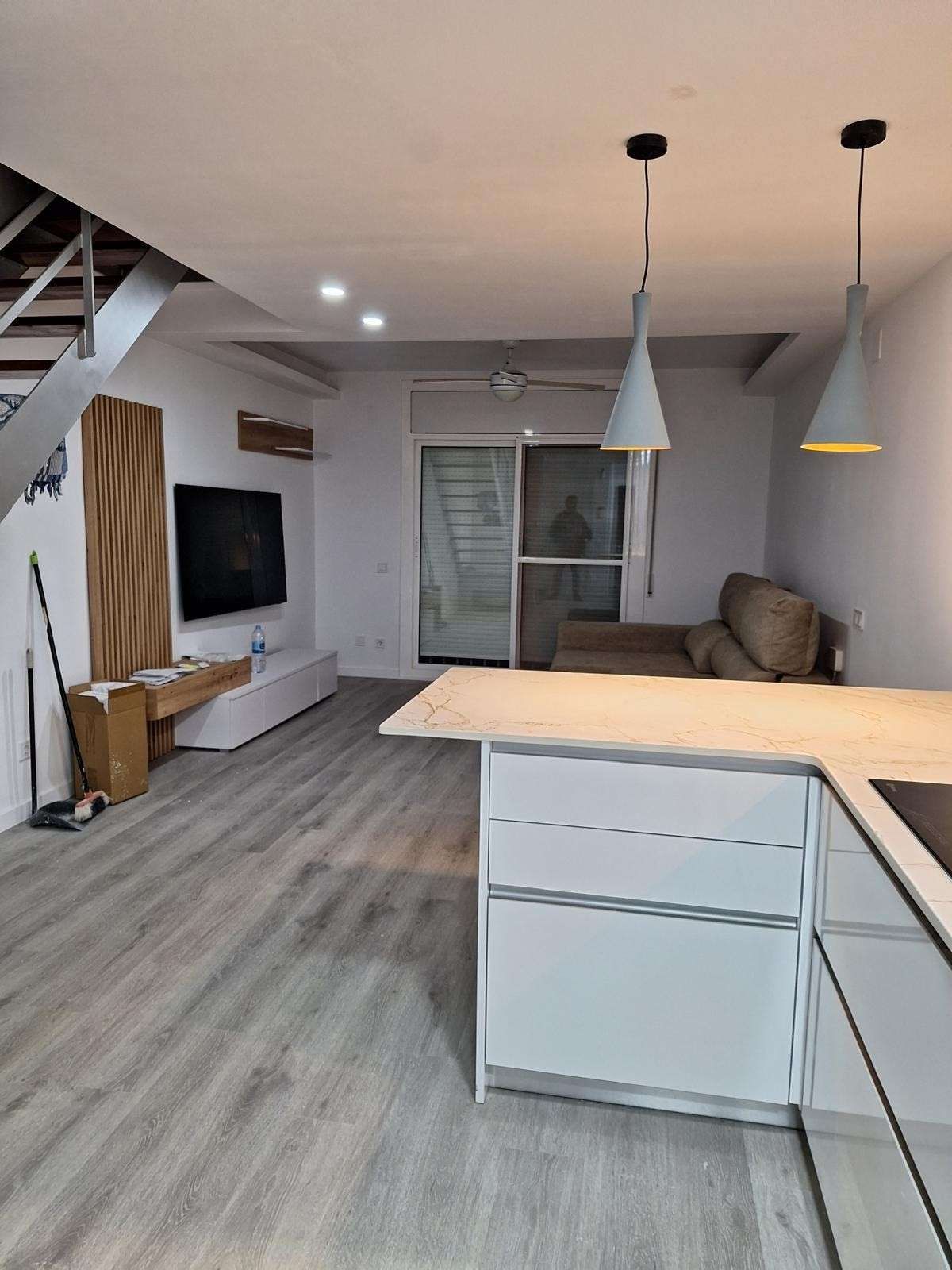 Salón moderno de planta abierta con suelos grises, muebles blancos, televisor, sofá y lámparas colgantes.