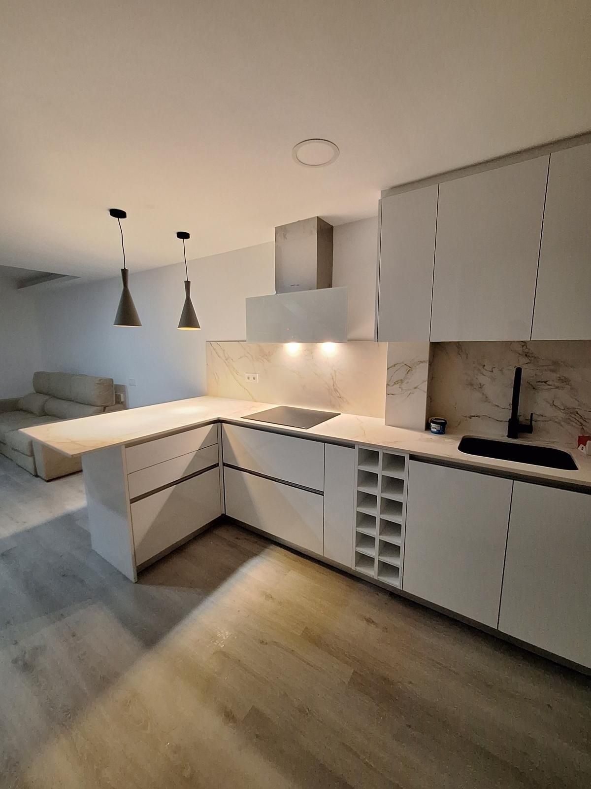 Cocina moderna blanca con encimeras en forma de L, lámparas colgantes, suelo de madera y electrodomésticos integrados.
