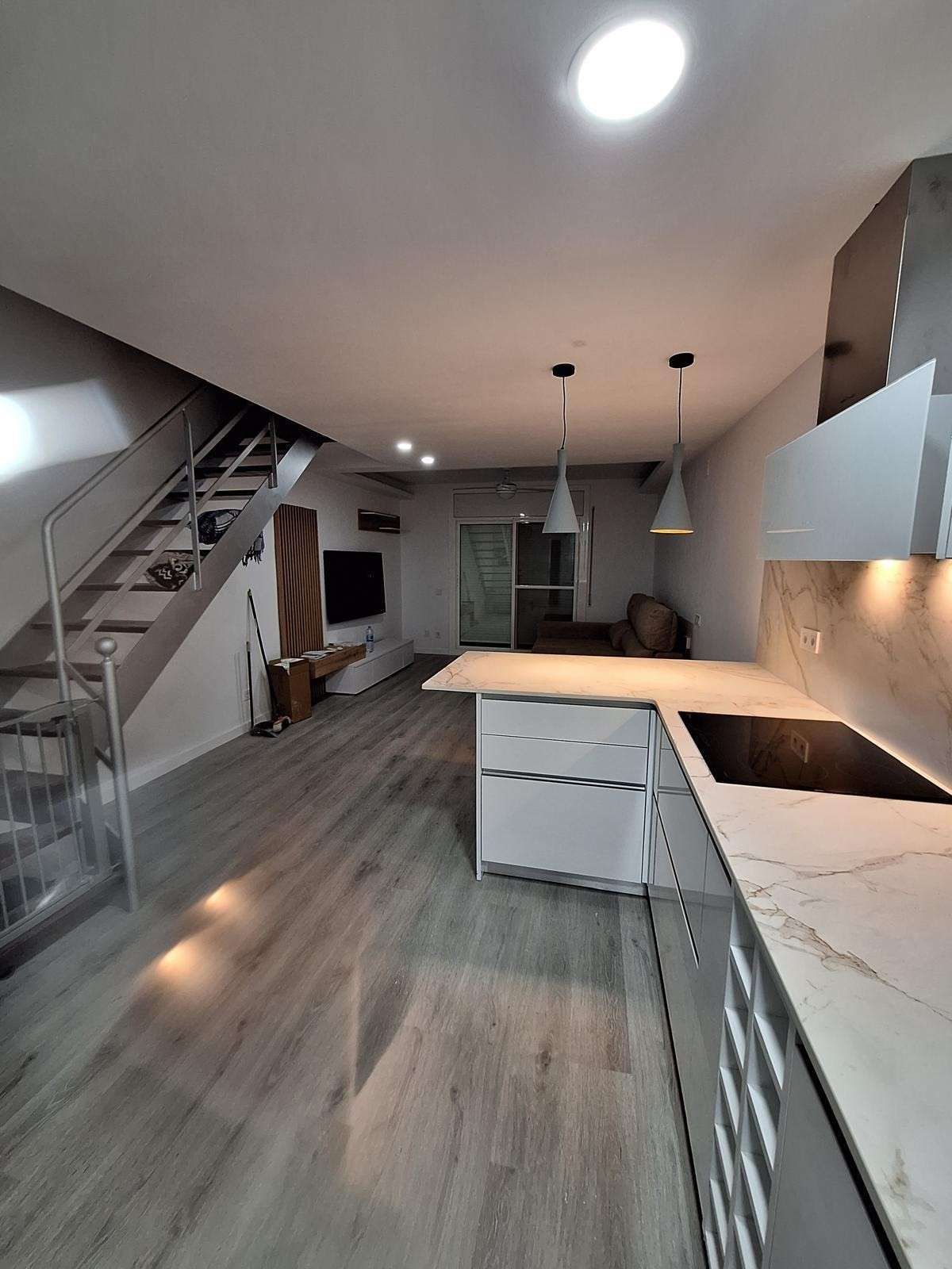 Cocina moderna con isla, gabinetes blancos, pisos grises, lámparas colgantes y escalera a la izquierda.