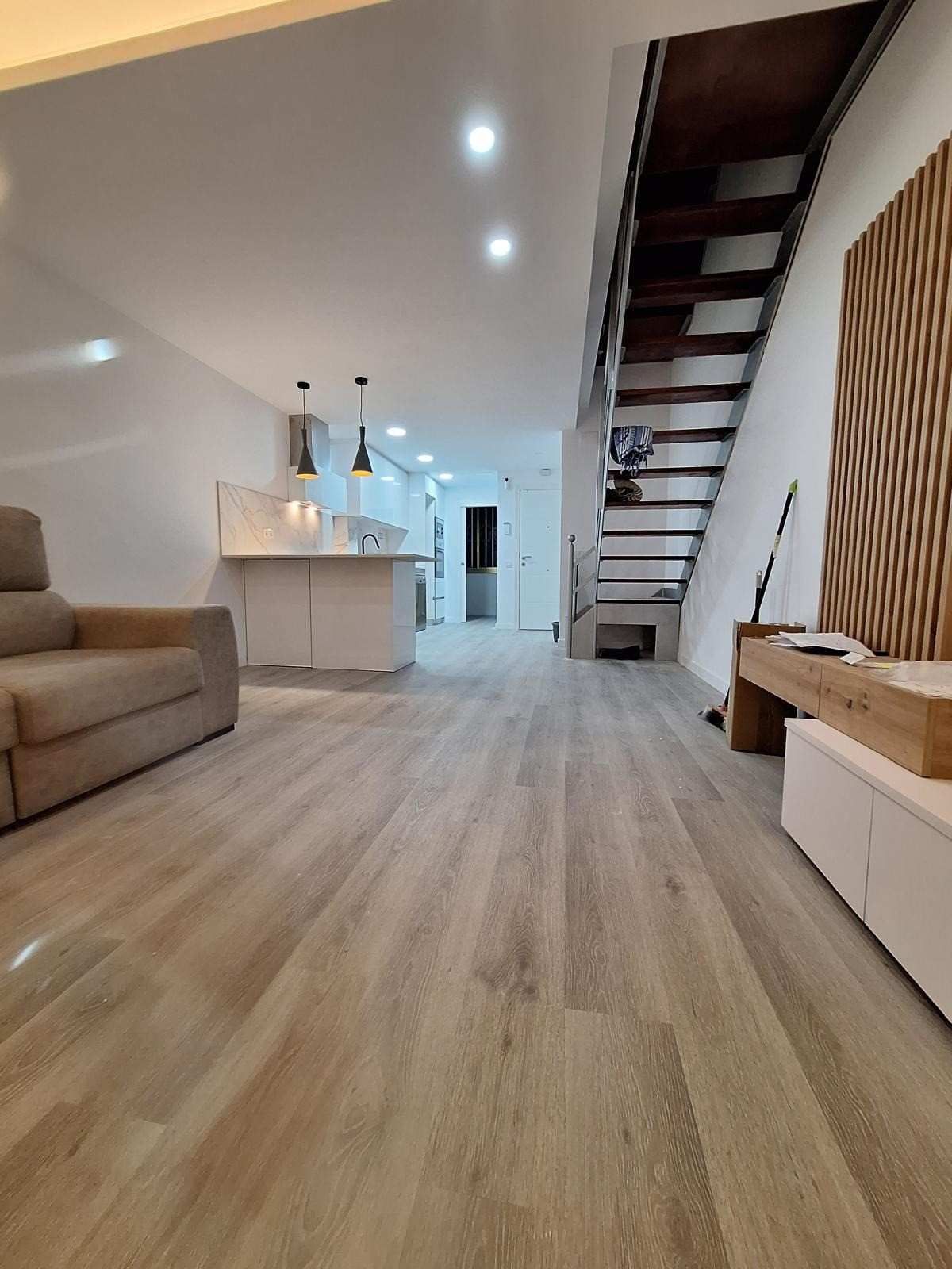 Salón moderno de planta abierta con sofá beige, suelo de madera, isla de cocina y escalera al altillo.