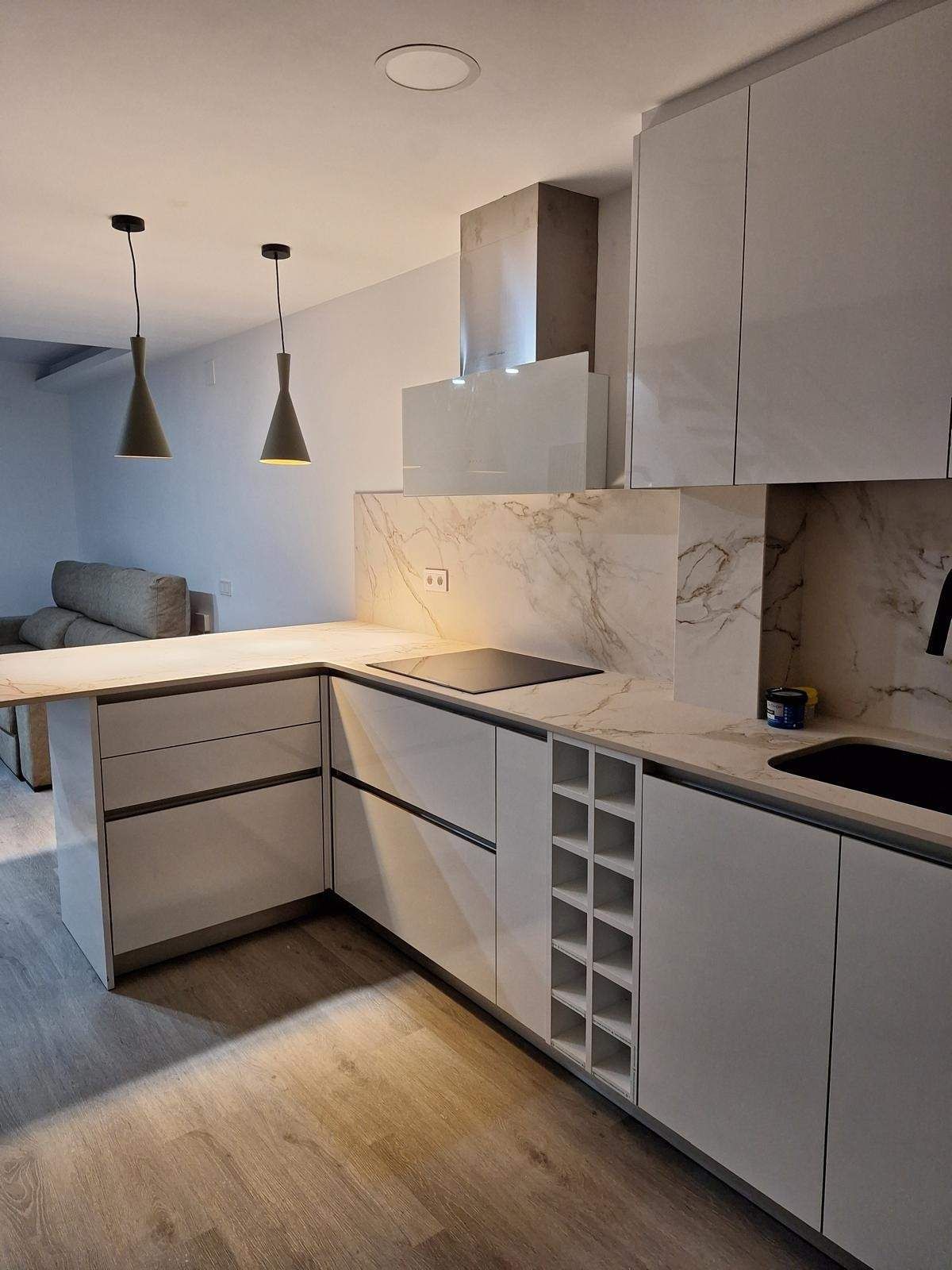 Cocina moderna blanca con salpicadero de mármol, isla central, lámparas colgantes y botellero empotrado.
