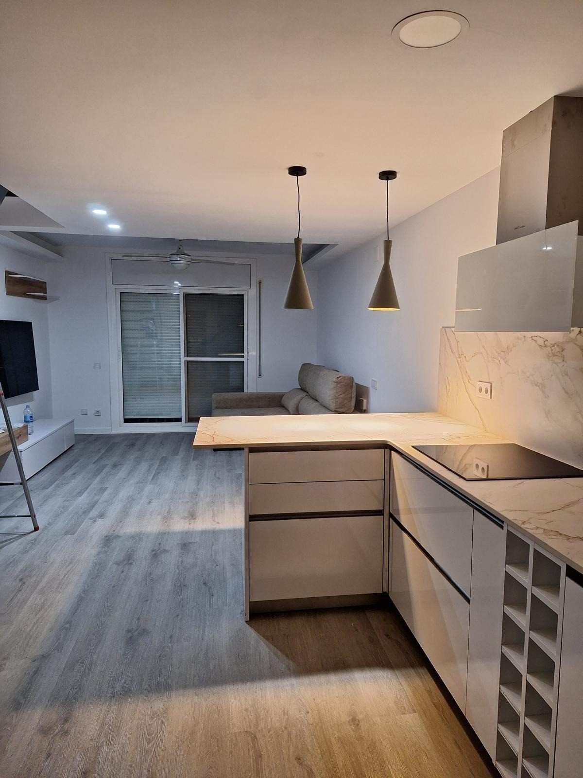 Cocina moderna con isla, lámparas colgantes, gabinetes grises y sala de estar abierta con puerta al balcón.