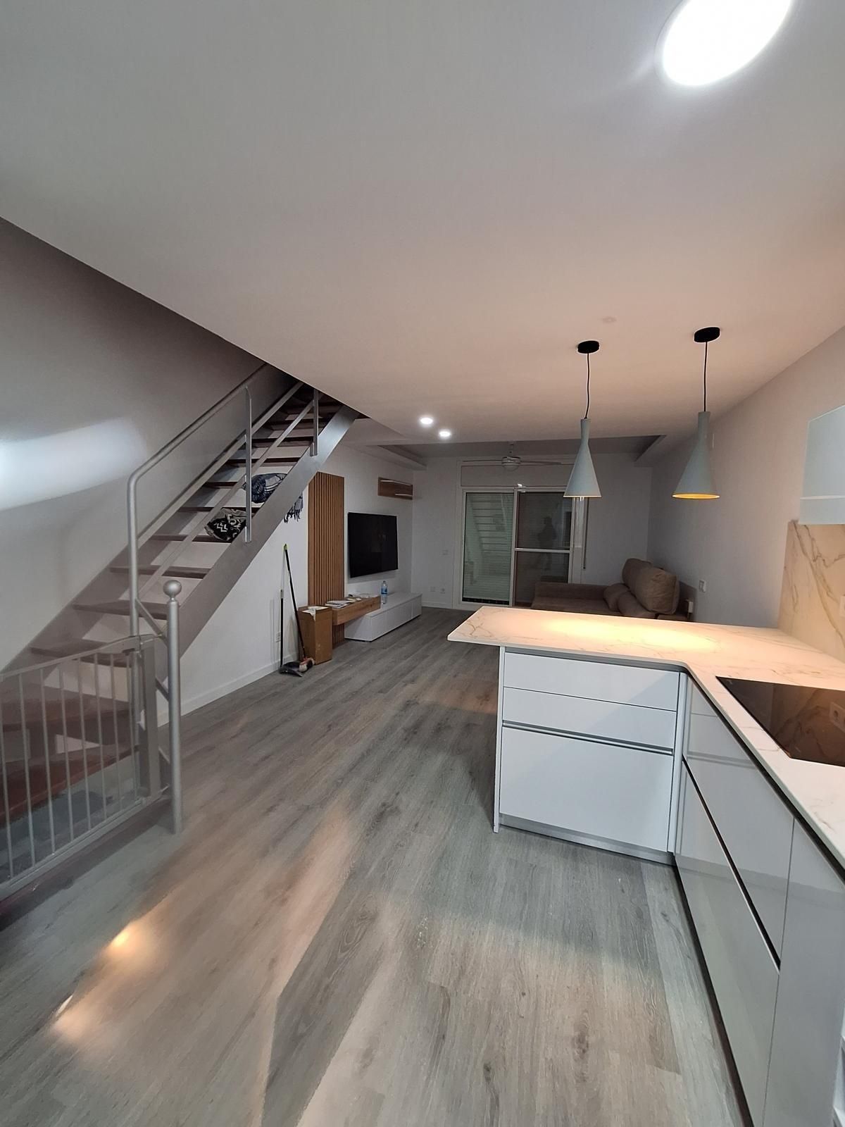 Cocina y sala de estar modernas de planta abierta con suelos grises, armarios blancos, escalera y lámparas colgantes.