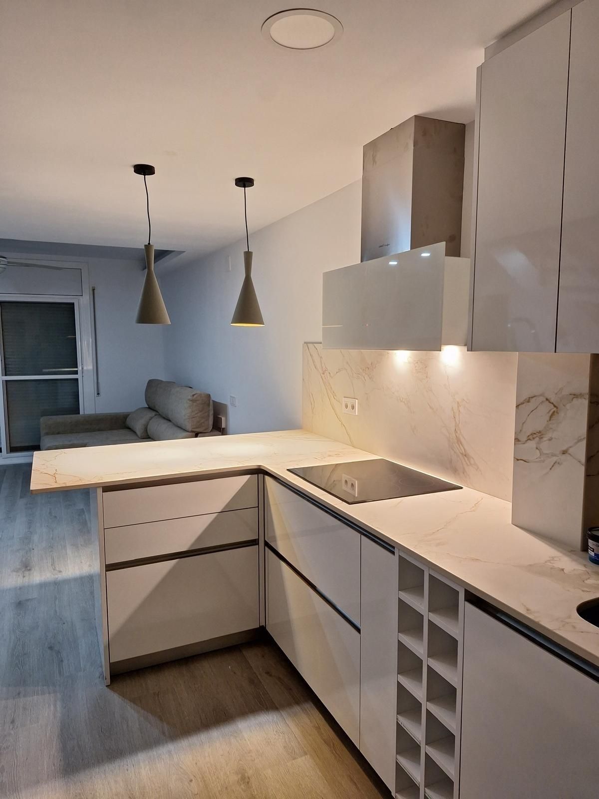 Cocina moderna blanca con encimeras de mármol, lámparas colgantes y vista abierta a un sofá gris en la sala de estar.