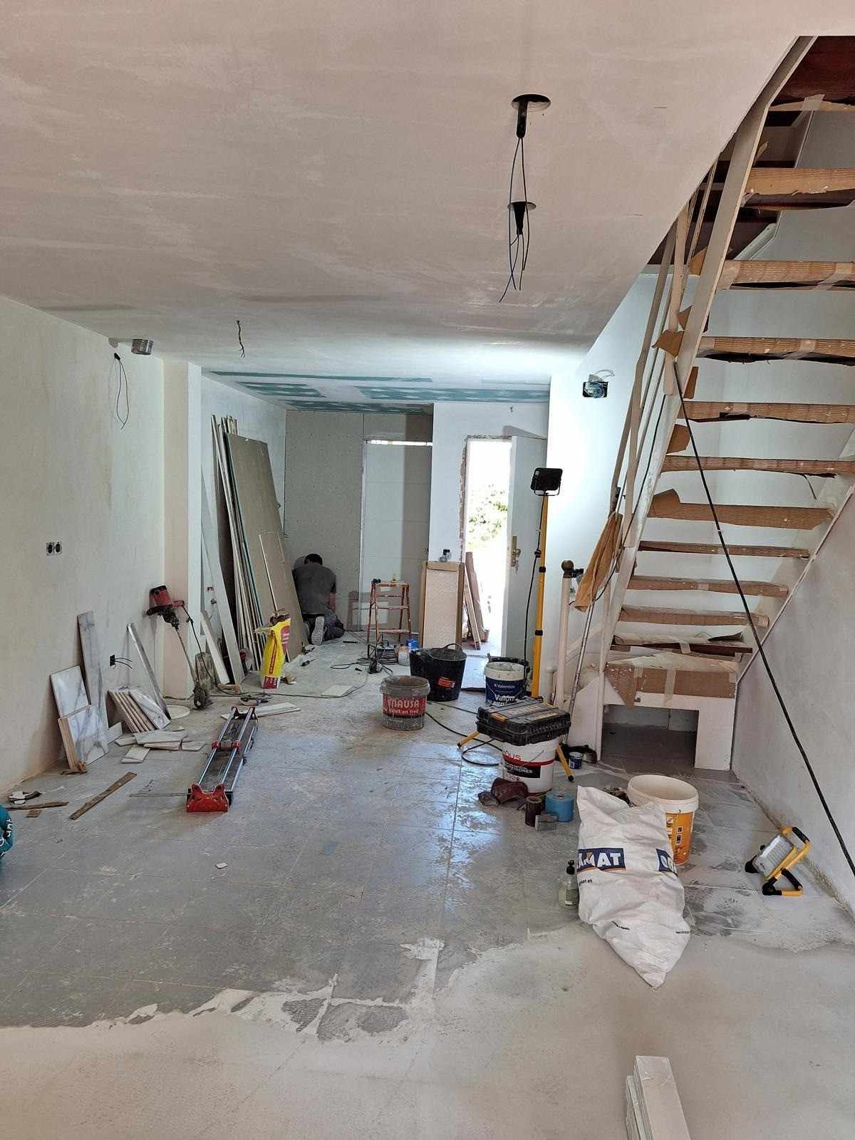 Obras de renovación en curso en una habitación estrecha con suelo sin terminar, escaleras a la derecha, herramientas y materiales esparcidos por el suelo.