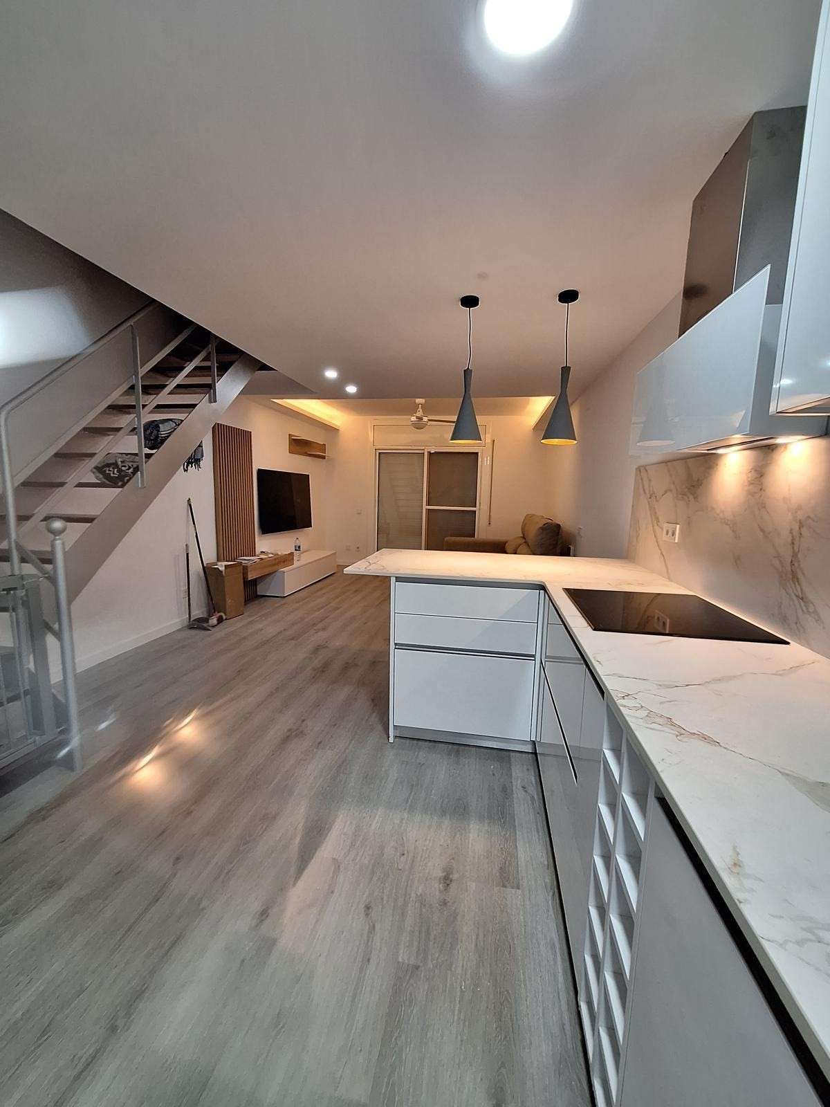 Cocina moderna con gabinetes blancos, encimeras de mármol, isla y escaleras en un espacio abierto.