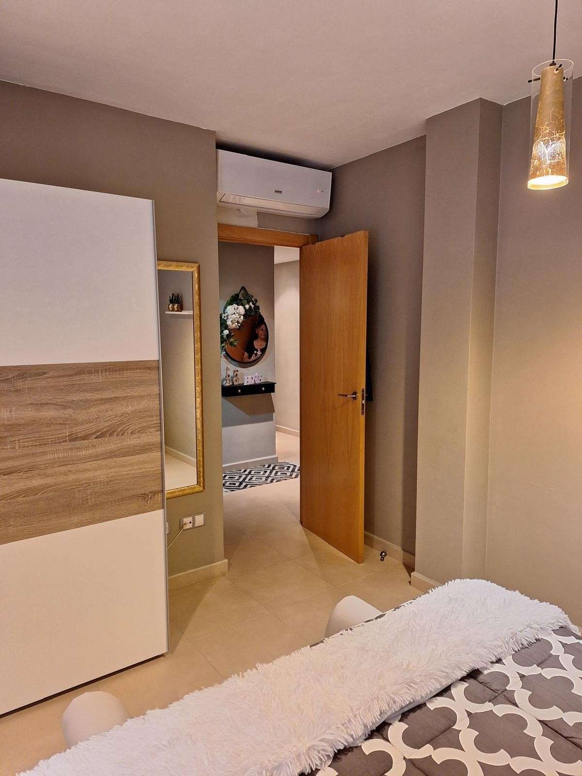 Dormitorio moderno con armario, puerta abierta, aire acondicionado mural y lámpara colgante en tonos neutros cálidos.