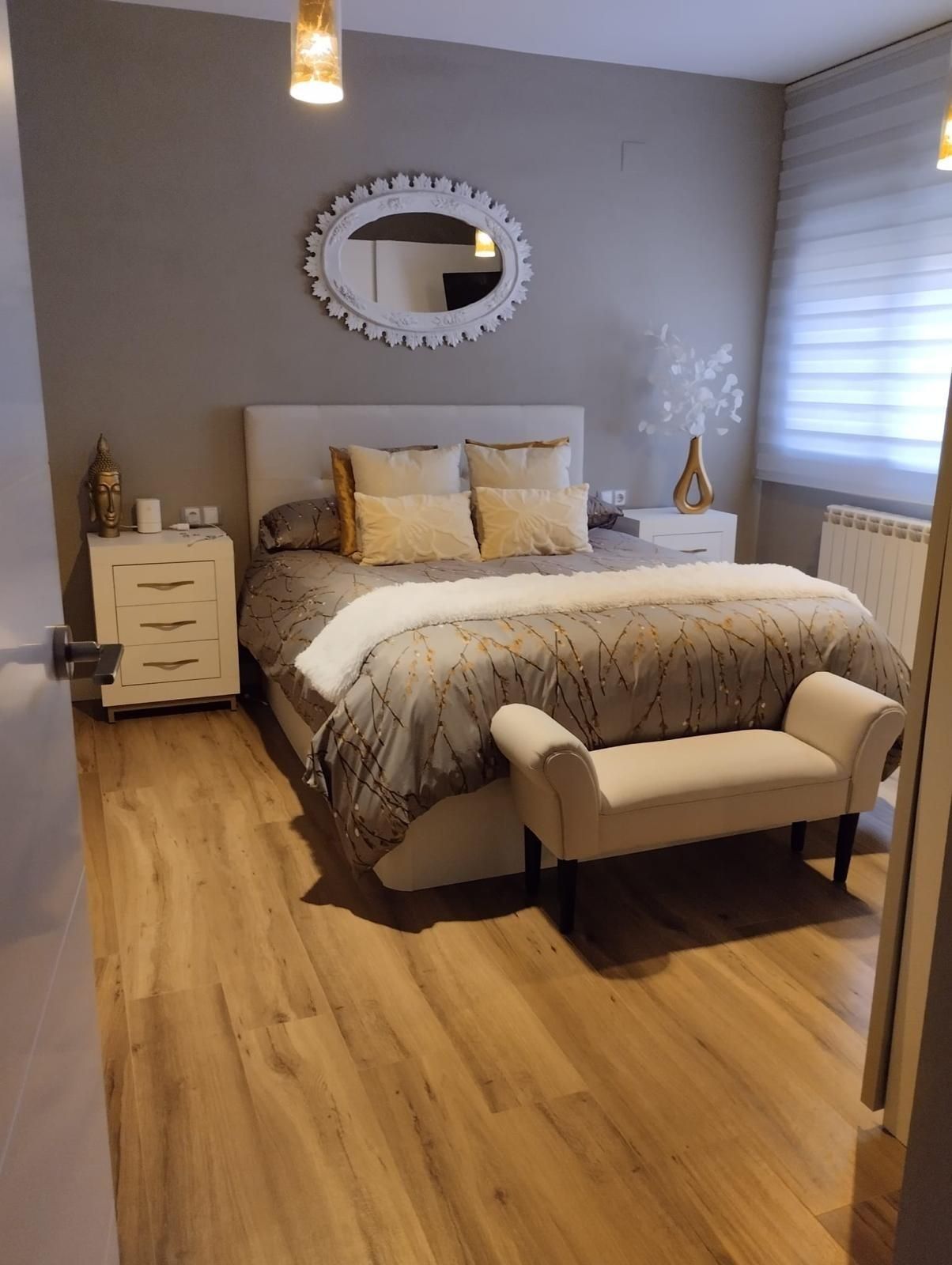 Acogedora habitación con paredes grises, suelo de madera, cama blanca, banco, mesitas de noche y espejo ovalado.
