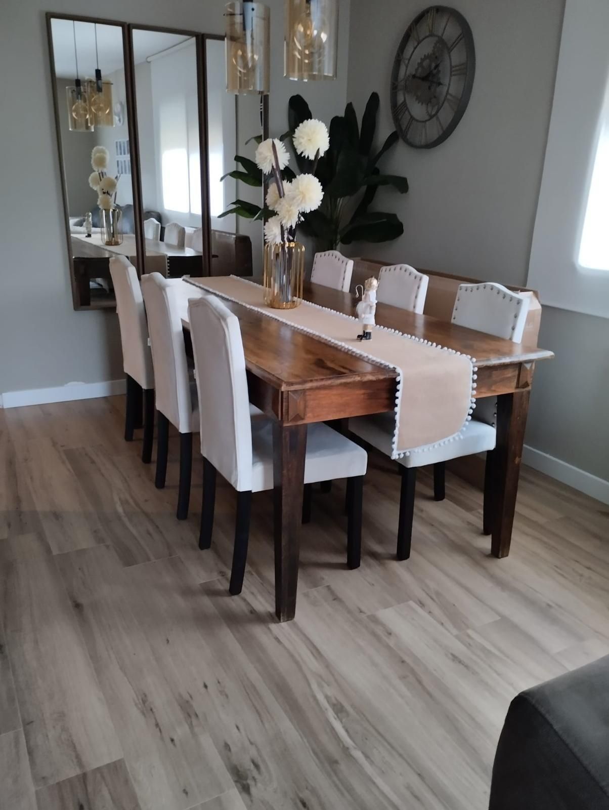 Comedor moderno con mesa de madera, sillas blancas, espejo y lámparas colgantes.