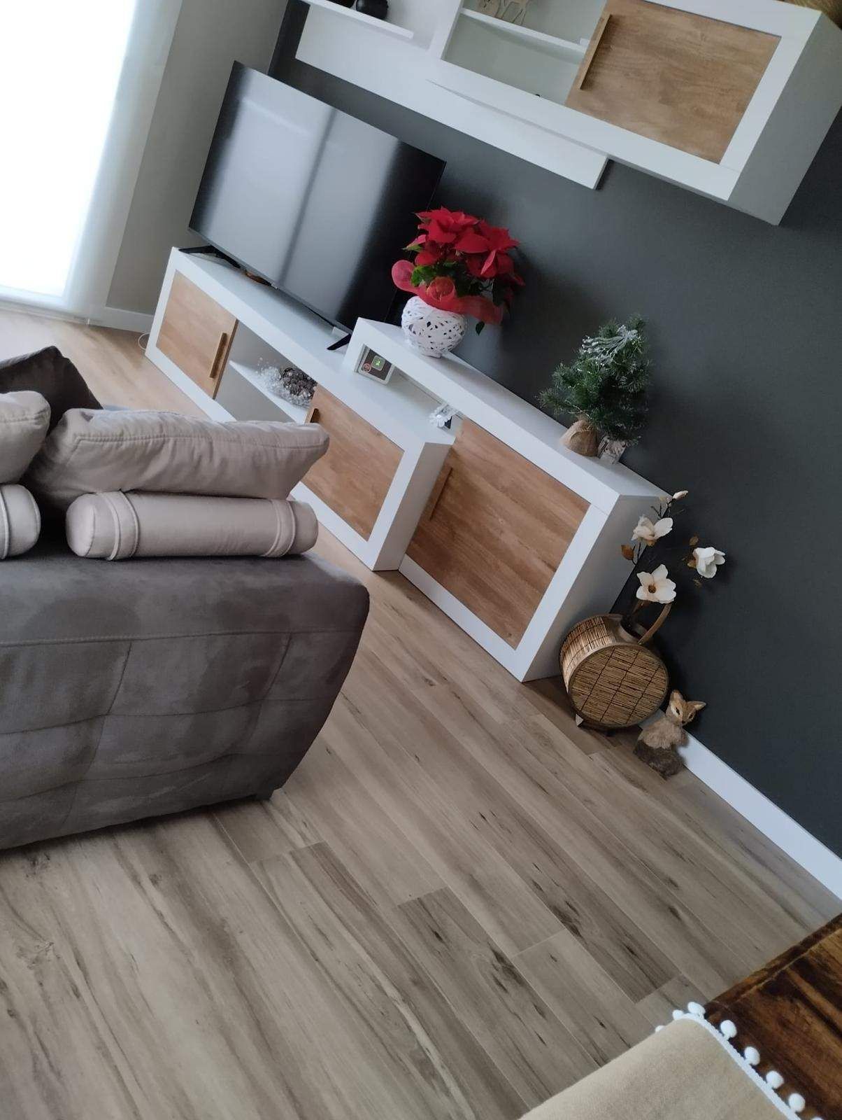 Sala de estar moderna con sofá modular gris, televisor montado en la pared, gabinetes blancos y piso de madera.